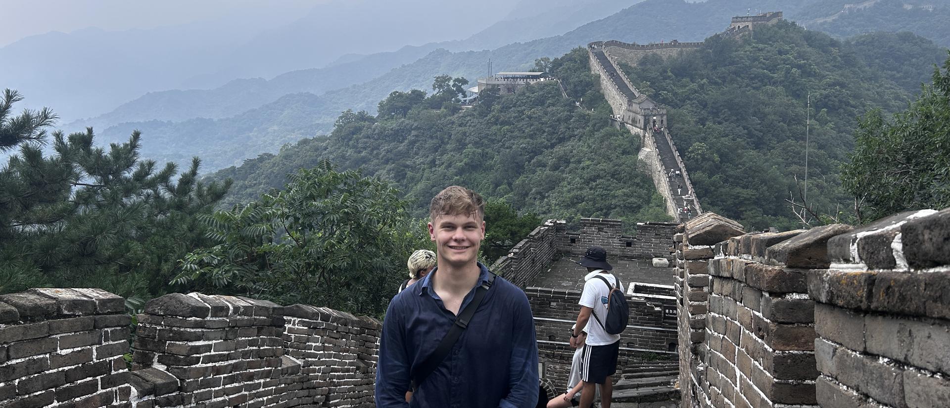 A smiling student stands on the Great Wall of China with misty mountains in the background.