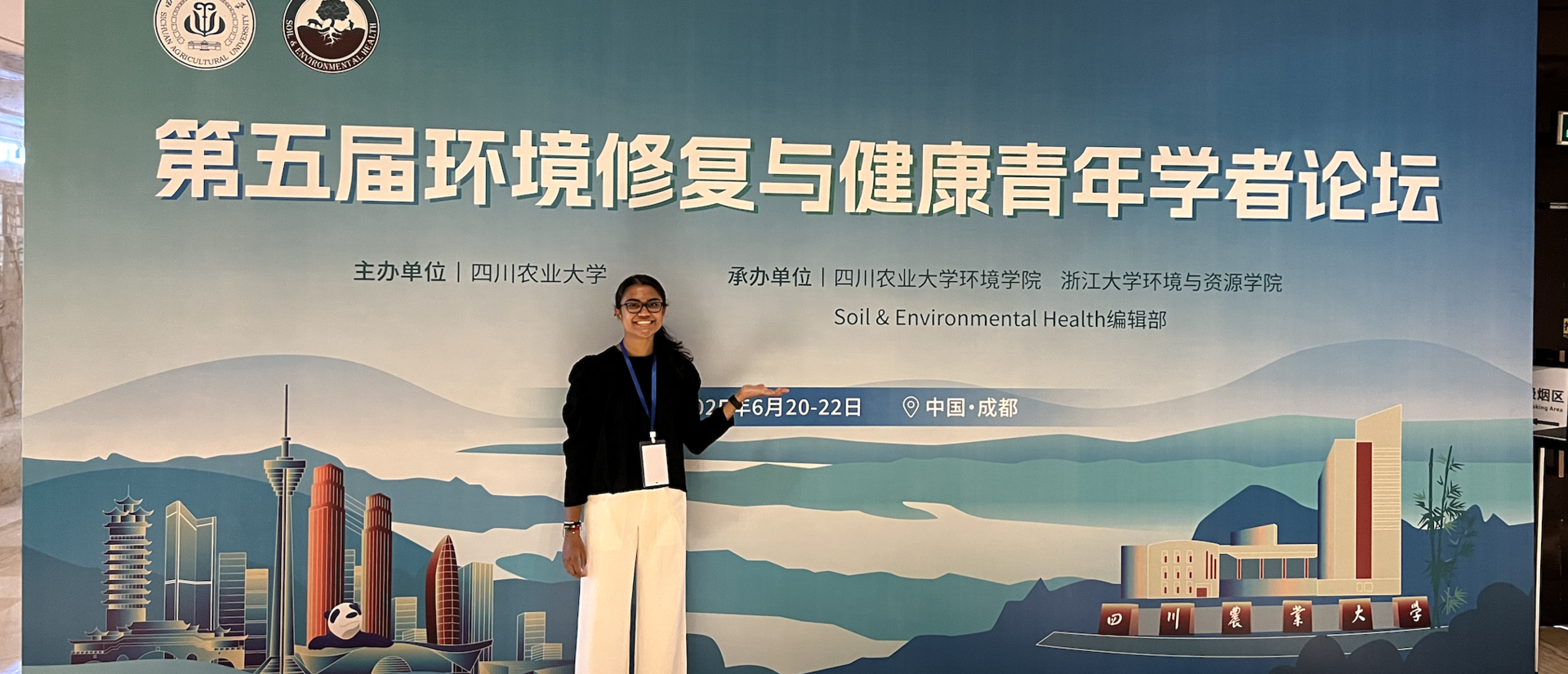 A student standing and smiling in front of a conference banner for the Soil and Environmental Health Forum in Chengdu, China.