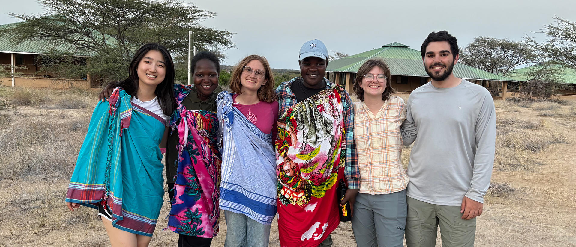 Group of six people smiling outdoors, some wearing colorful patterned wraps, with dry grassland and buildings in the background.