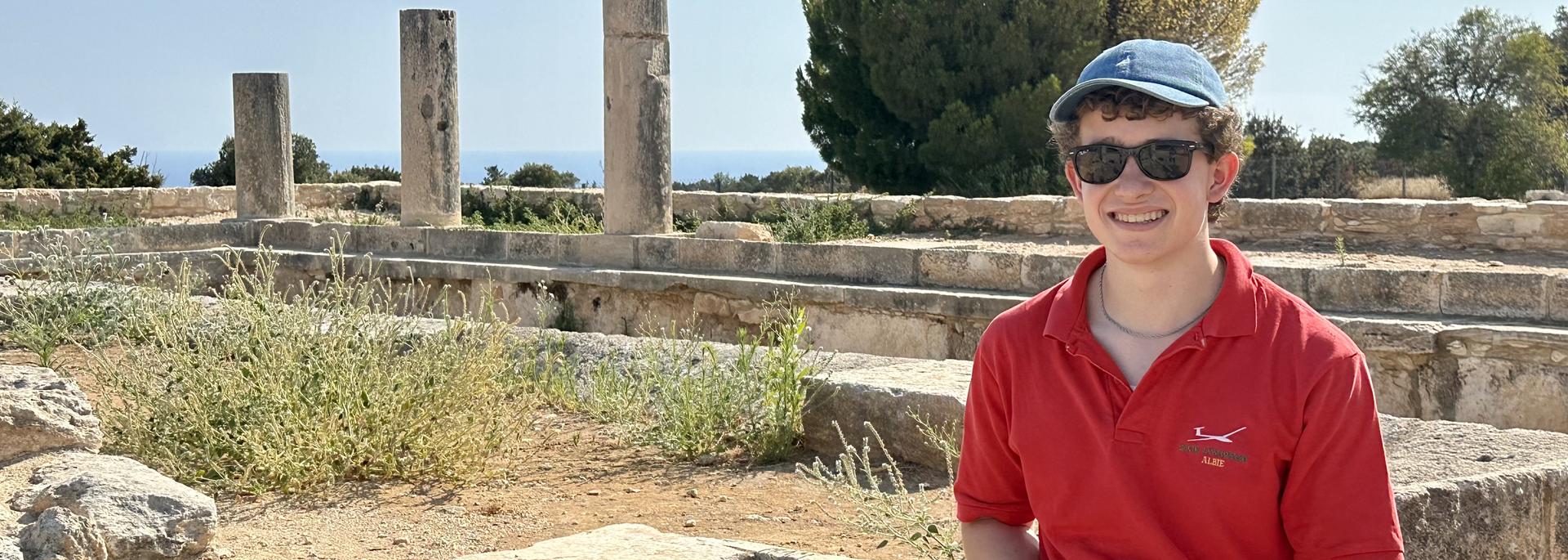 A smiling student in a red shirt and blue cap sits among ancient stone ruins with columns and trees in the background.
